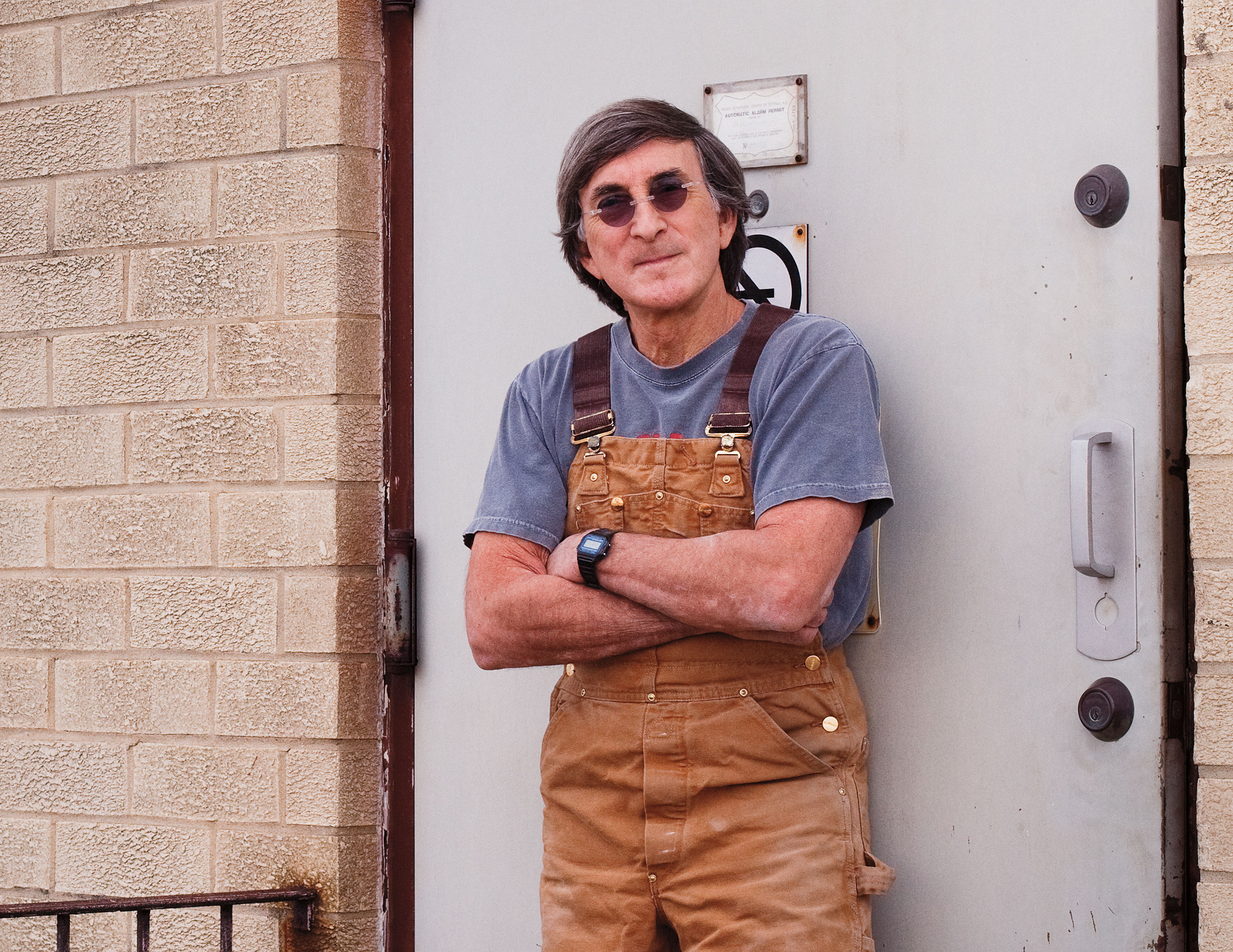 A man wearing sunglasses, a blue T-shirt, and brown overalls leans against a door