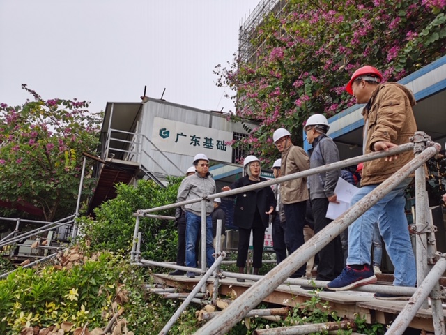 A woman with gray hair and a group of middle-aged men, all wearing hard hats, stand on a platform outside of a building that's under construction. 