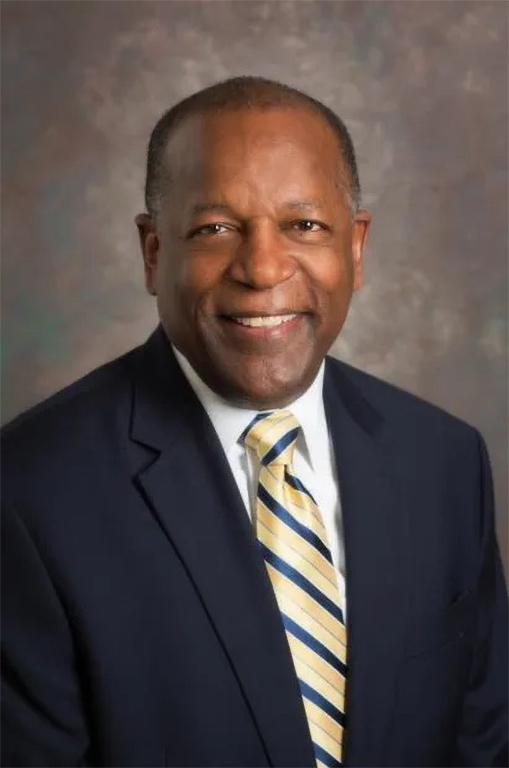 Professional photo of a Black man dressed in a dark blue suit jacket, a yellow and blue striped tie, and a white button-up dress shirt. 