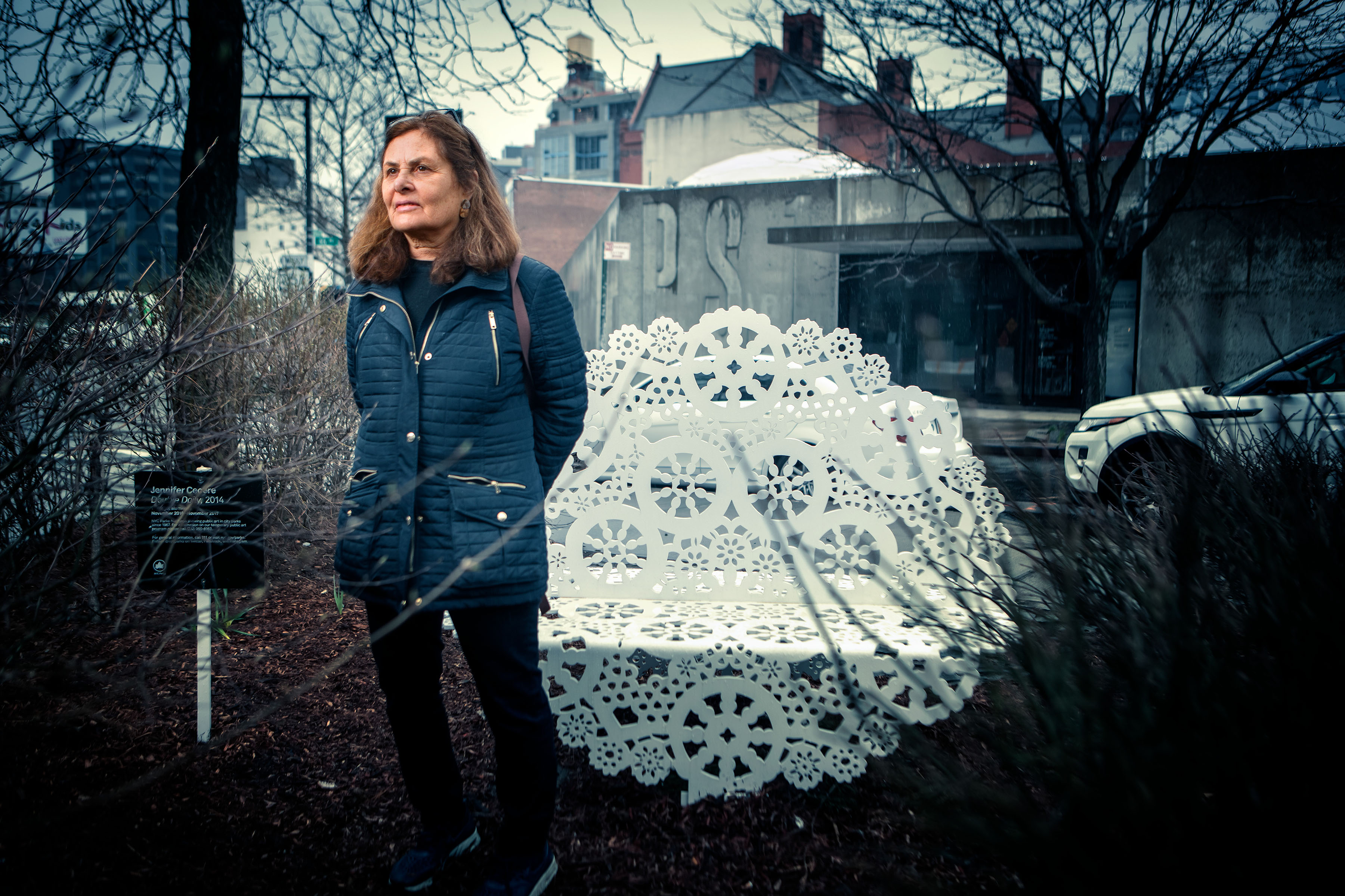 A woman with shoulder-length brown hair, dressed in a blue coat, stands in front of a large white doily sculpture outside.