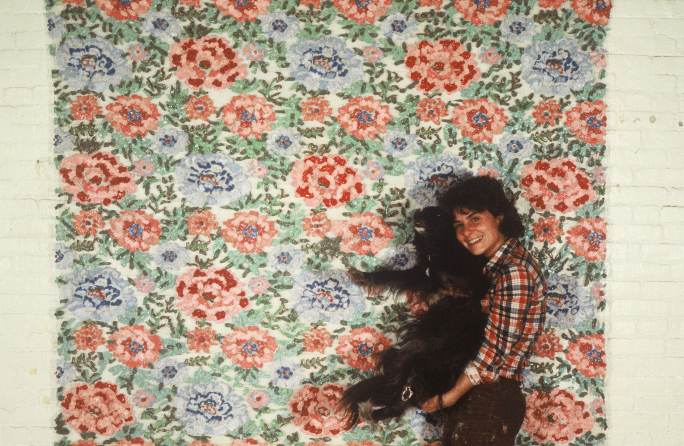 A woman holding a dog in front of a flowered backdrop covering a brick wall.