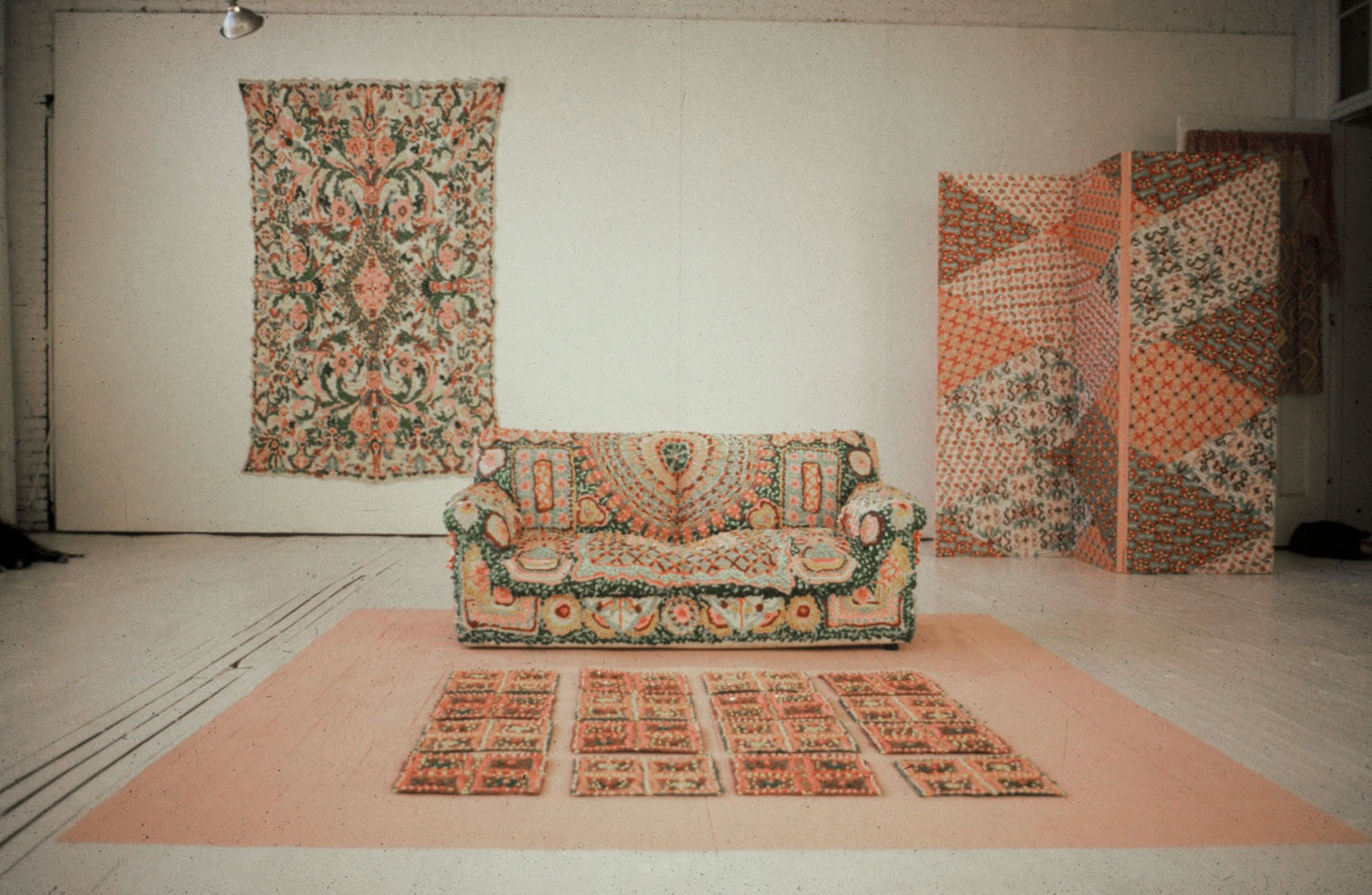 Front-facing view of an installation featuring a colorful couch patterned with doily-like motifs, a rug with a quilt-like design, a colorful room divider, and a colorful rectangular wall hanging.