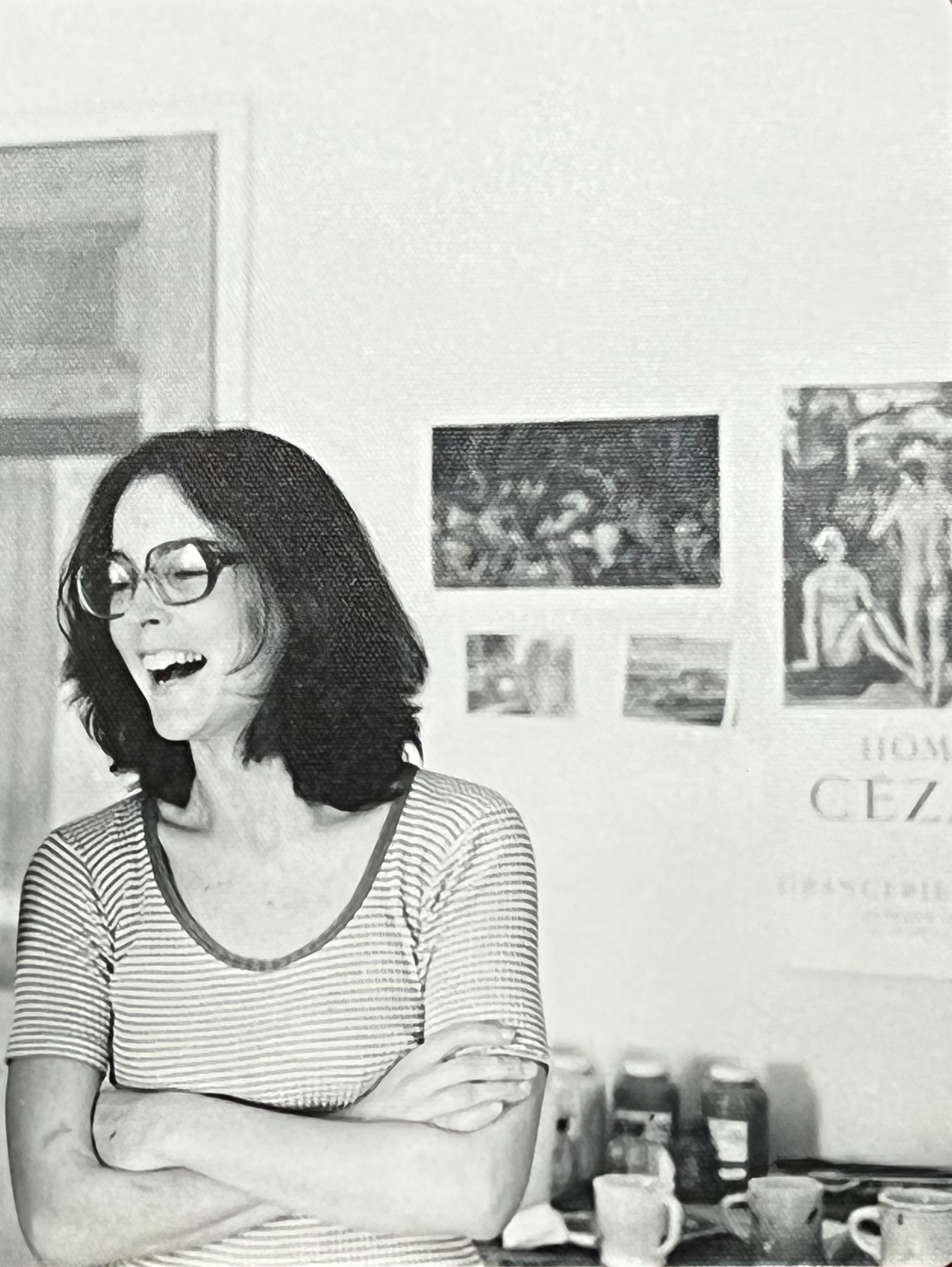 Black-and-white photo of a woman laughing in a room with art prints on the wall behind her.