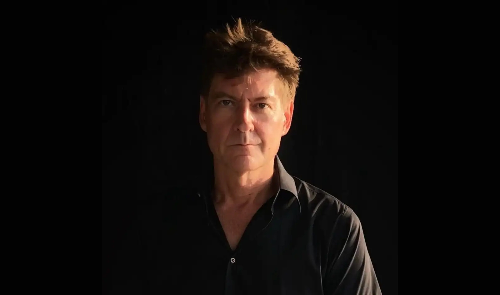 Portrait photo of a white man with brown hair, dressed in a black button-up shirt, against a black background.
