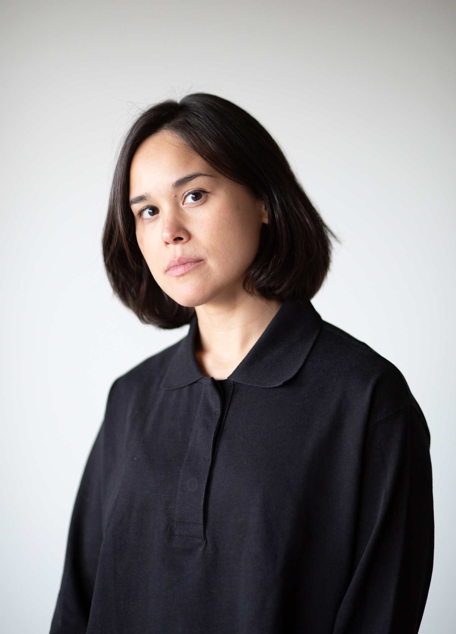 A woman, dressed professionally in a black collared shirt, stands in front of a white wall and looks at the camera.