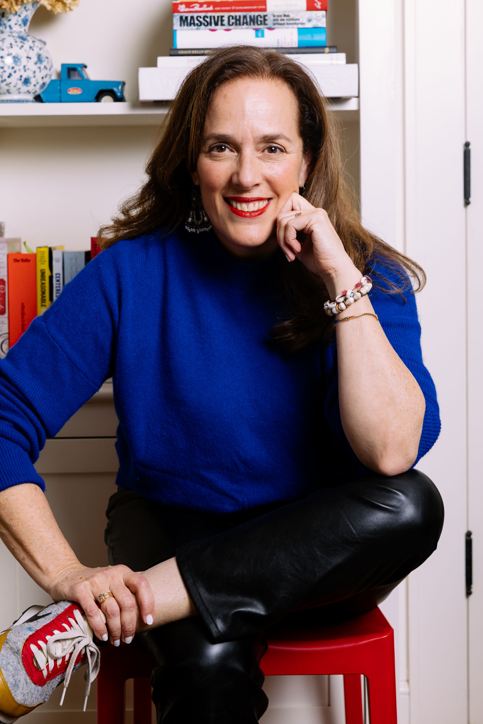 A woman with long brown hair, dressed in a vibrant blue sweater, black pants, and red-and-gray sneakers, sits on a red stool