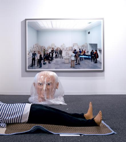 image of a Maurizio Cattelan piece of Picasso figure wrapped, the head is wrapped in plastic and the body is laid on a moving blanket with a  photograph in the background