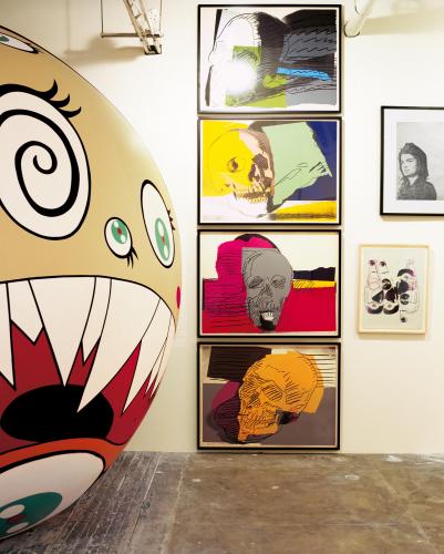 A view of many pieces of art, a Takashi Murakami on the left, a large inflatable work of a character with wild eyes and a mouth, and a stacking of Warhols with more images and artworks to the right