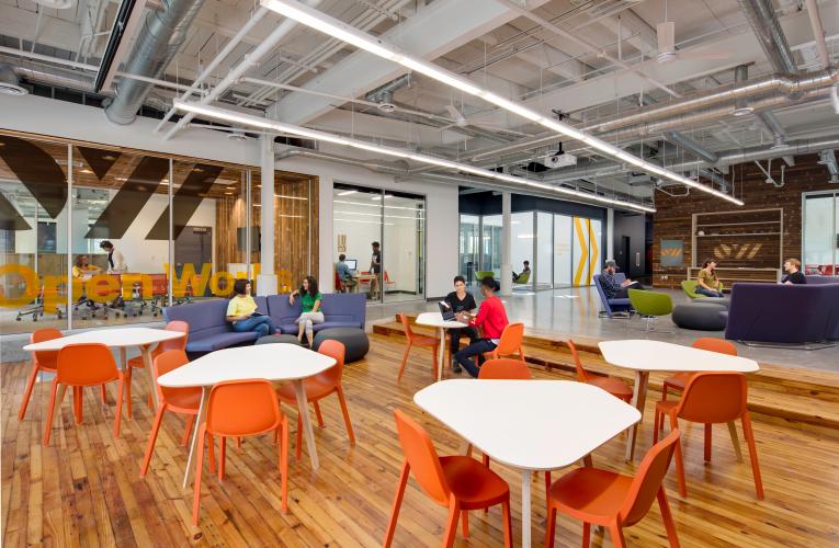 Building lobby with wooden floor, white tables, and orange chairs.