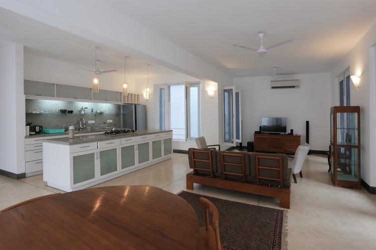 An apartment contains wooden furniture in an open living room and white drawers and frosted glass cupboards in an open kitchen.