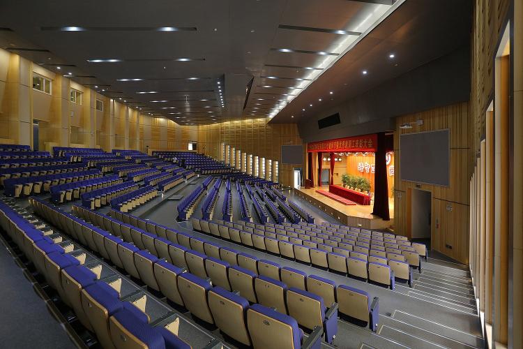 Blue seats in a large, empty auditorium face a lit stage.