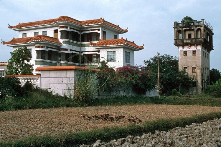 A newer white building with a red roof stands in contrast to an older tower near the entrance to a village.