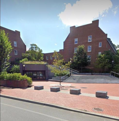 A single-story entrance leads into a large, multistory brick building. The sidewalk is also made of bricks. Cement stairs wrap around the the entrance and lead up into a courtyard outside the brick building.