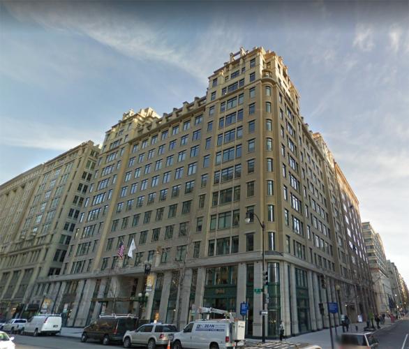 People cross the street in front of a tall, brown building.