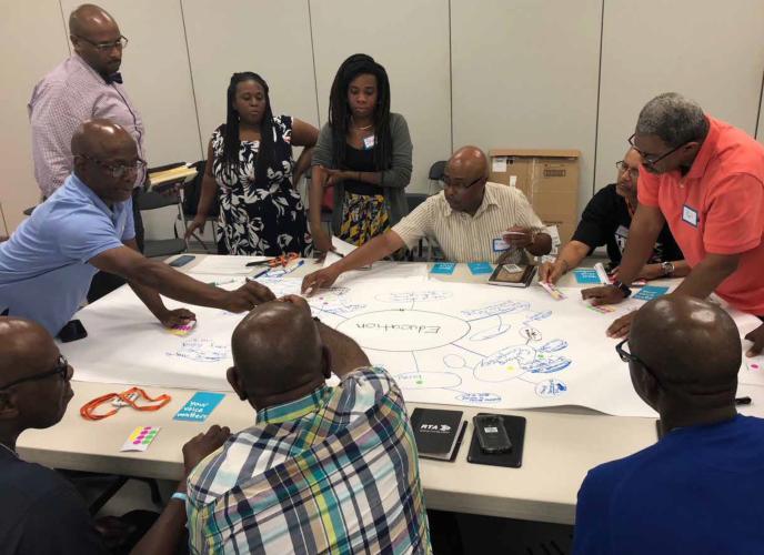 A group of adult Black men and two Black women are gathered around a table as they create a diagram on a large sheet of paper.