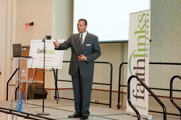 A professionally dressed black man delivers a speech from a small stage.