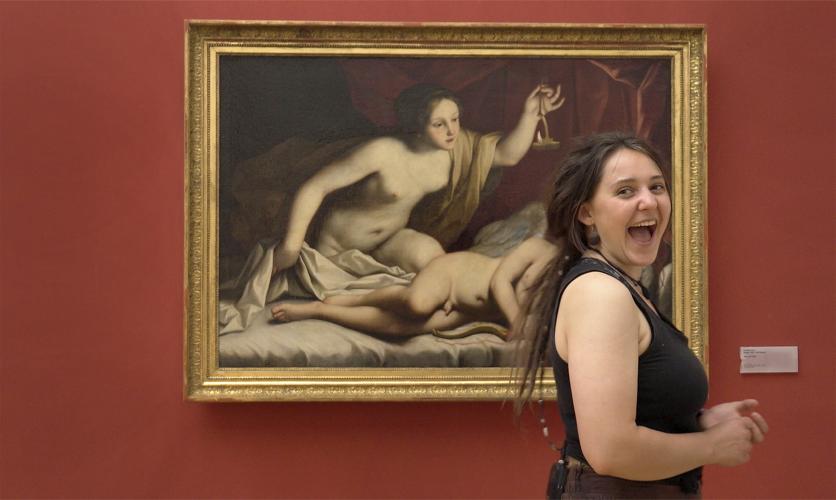 A teen girl laughs in front of a painting of a nude woman and nude angel in a bed.