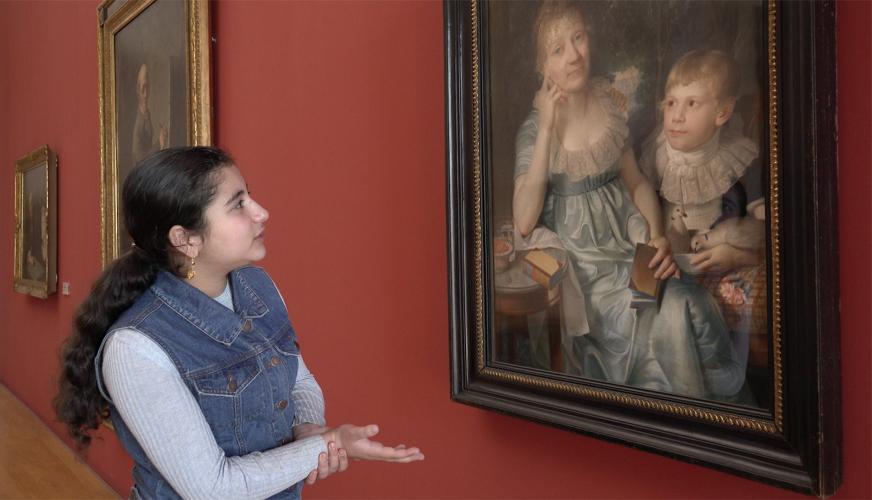 A teenage girl looks upon a painting of an elderly woman and an adolescent boy.