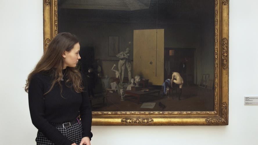 A teenage girl dressed in a long-sleeve black shirt looks at a painting of a woman dressing in a large room. 