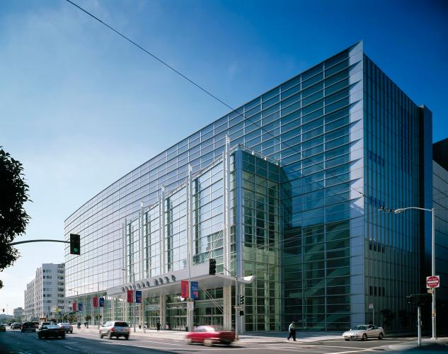 Cars drive past a multistory, glass building on a blue-sky day.