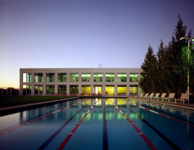 A long, multilane pool leads toward a two-story building lined with two rows of large windows lit up against a purple  dusk sky.