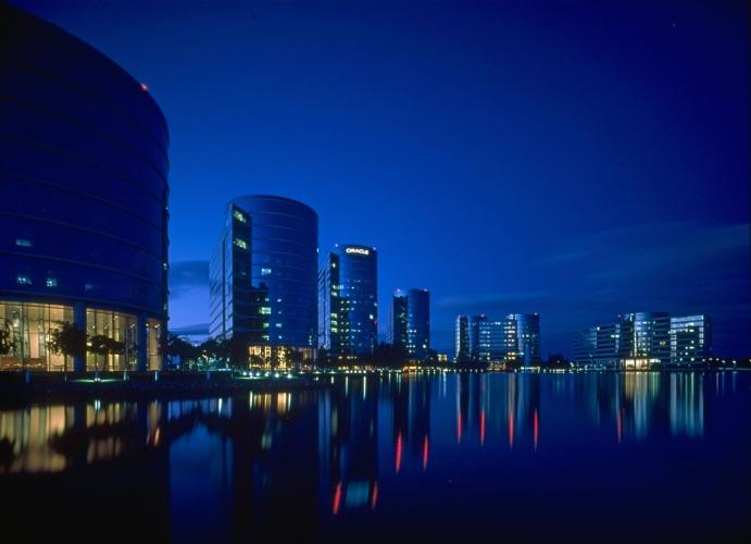 Six buildings tower over a pool at night, reflecting light shining from the building lobbies. 