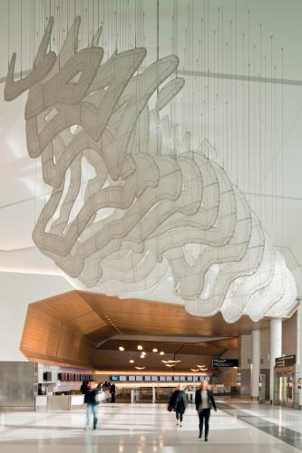 An airport terminal features a suspended sculptural artwork made from powder-coated steel tubing and greenhouse shade cloth.