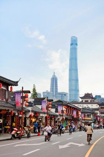 A skyscraper stands tall in the distance, overlooking traditional Chinese buildings decorated with paper lanterns as people ride bicycles in the street.