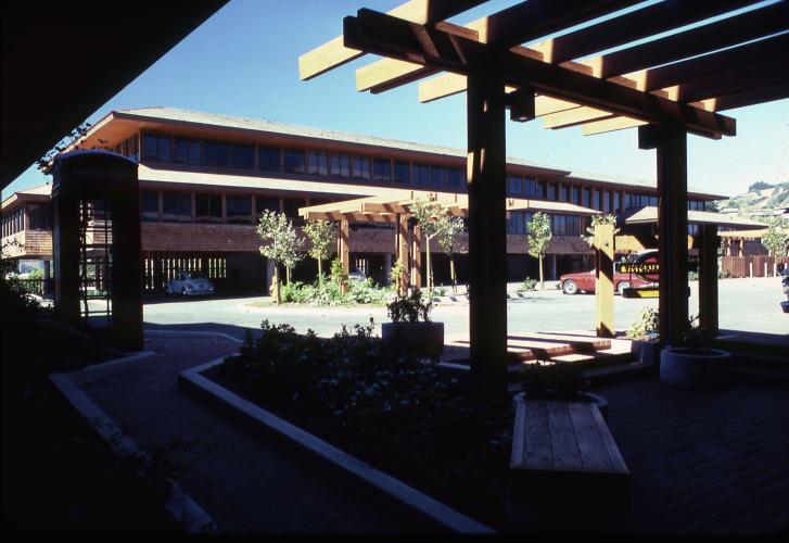 Two wooden pergolas and small trees stand in front a multistory building lined with windows.