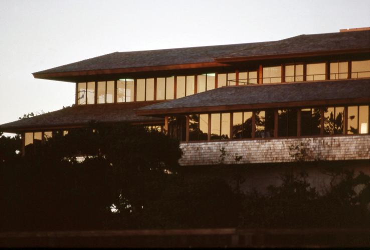 A multistory lined with windows reflects the surrounding trees at sunset.