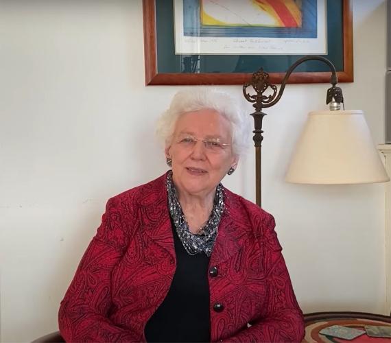 An older woman with white hair, wearing a red jacket over a black shirt, looks at the camera for a video interview.