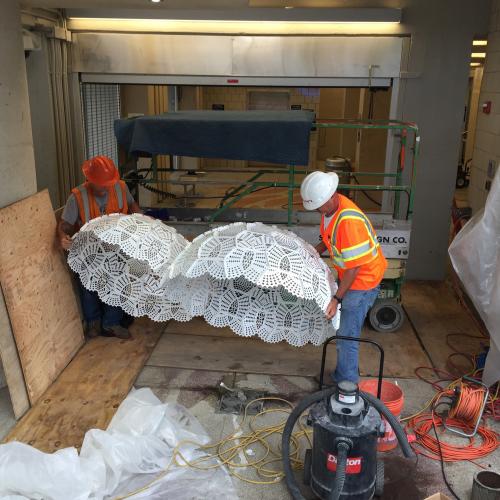 Two construction workers carry a large sculpture of two large white doilies.