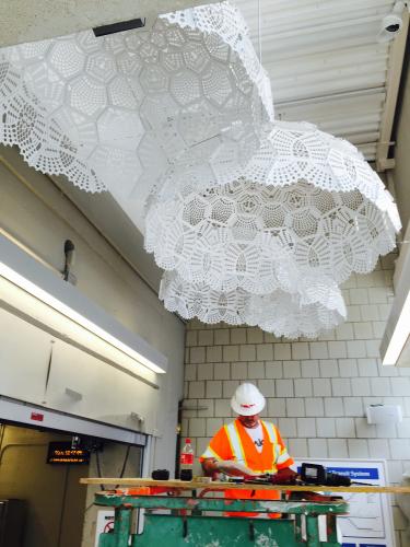 Four large white doily sculptures hang from the ceiling over a man dressed in a white hat and an orange safety vest.