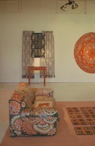 An installation view of a colorful couch patterned with doily-like motifs, a rug with a quilt-like design, a large doily wall hanging, and transparent gray curtains hanging on a wall.