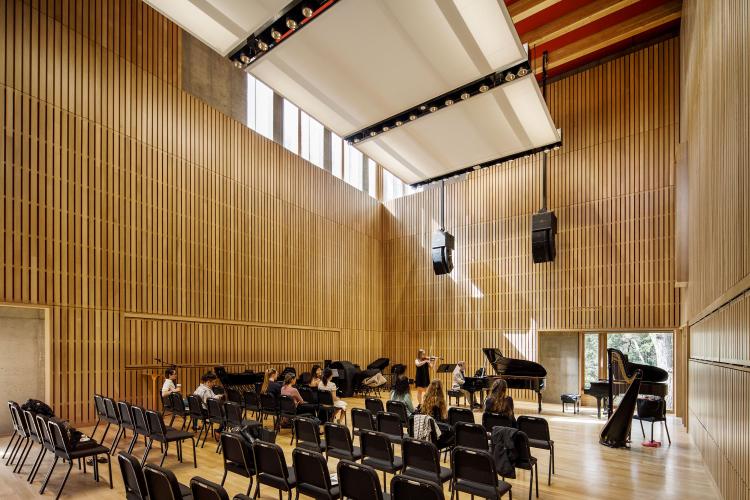 Vertical planks of wood line the walls of a large rehearsal hall where black chairs face the front of the space and students watch a professor and another student playing musical instruments.