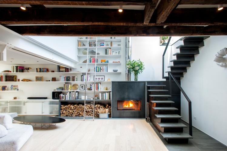 A spacious and airy living room featuring light-colored wood flooring, white walls, and a wall lined with white shelves contrasted with a black fireplace and black, open staircase.