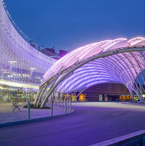 The glass curved facade of the steel grid-shell is lit up at night with a purple glow. 