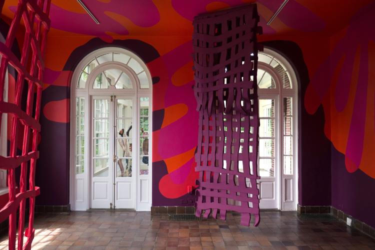 An installation art exhibit features orange, fuchsia, and dark purple abstract shapes painted on the walls and a purple grid-like tapestry hanging from the ceiling.