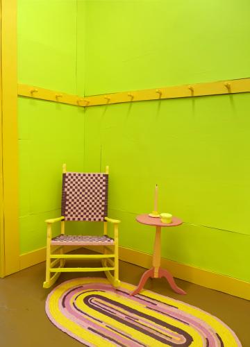 A room with lime green walls and yellow trim features a yellow and pink rocking chair, a coral-colored side table, and a round yellow and pink rug.
