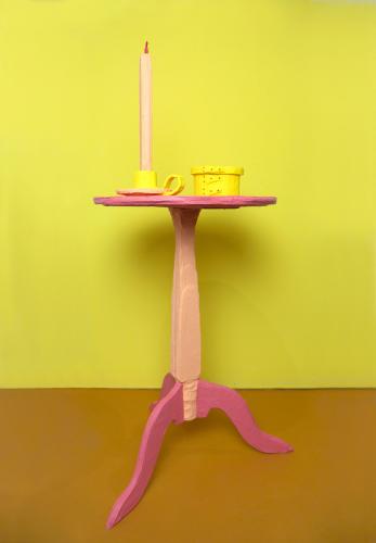 A pin candle in a yellow candle holder sits on a small pink side table in front of a yellow wall.