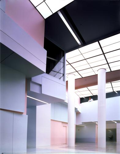 Photograph of a building interior with white walls and columns accented by pale red wall sections and lights set in a rectangular grid across the ceiling.