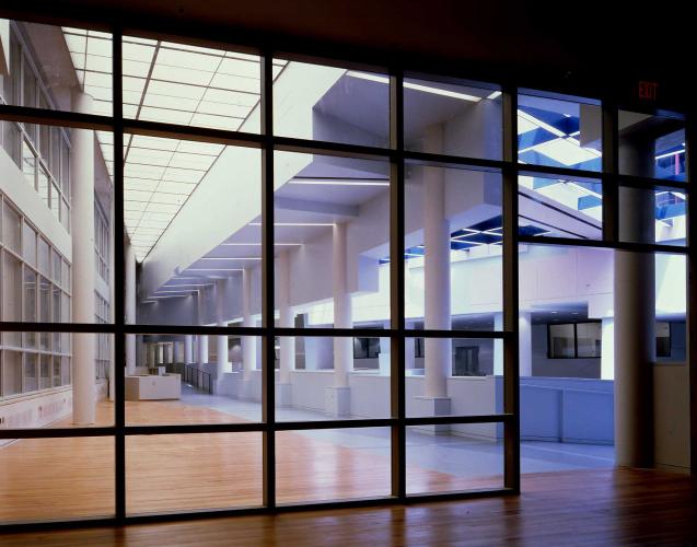 Photograph of a building interior, looking into a long hallway with white columns, from behind a large window subdivided into a grid with narrow partition.