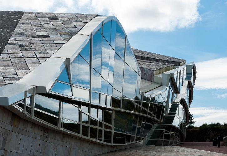 Detail view of curving building with wall of windows reflecting a bright blue sky and clouds.