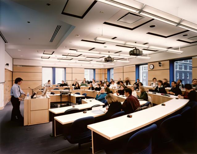 A professor lectures at the front of the room as students look on, seated in the lecture room.