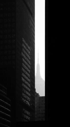 A skyscraper in the distance peeks through a crack between two buildings in the foreground in a black-and-white photo.