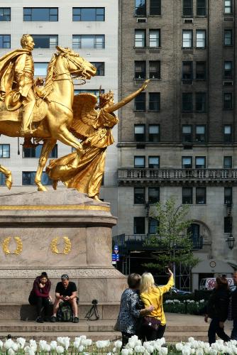 A couple takes a selfie photo, echoing the pose of a larger-than-life gold statue of a winged woman reaching her arm out toward the sky in the background.