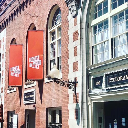 A two-story building with a red brick facade features arched windows on the second level and two red signs for the Boston Center for the Arts