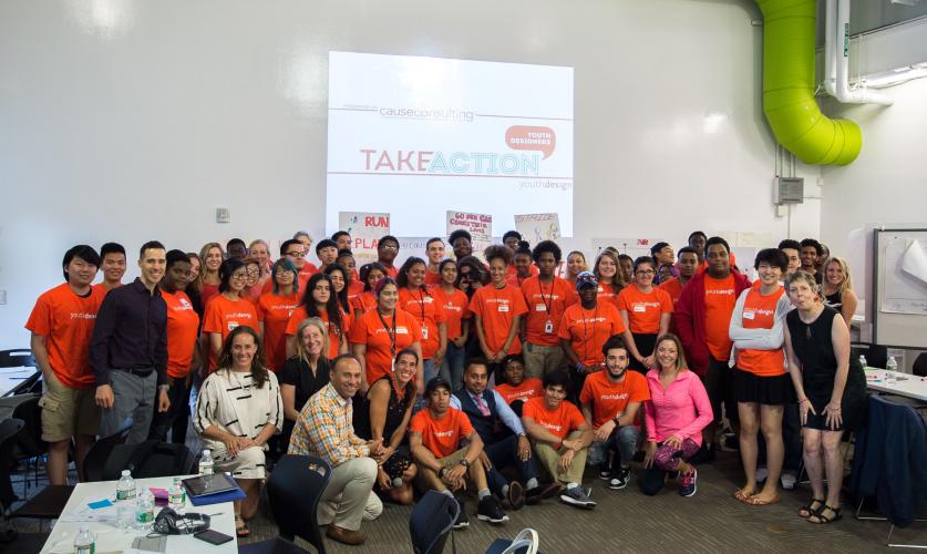 A large group of teenagers dressed in bright orange T-shirts pose for a photo with the mentors for a youth design workshop