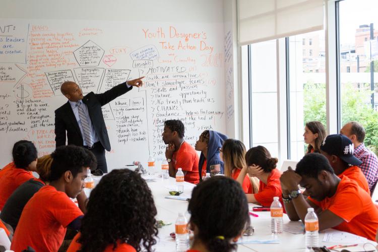 Teenagers dressed in orange T-shirts sit at a long table, while an adult male in a business suit lectures at the front of the room.