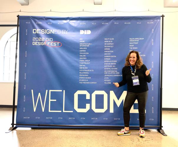 A woman with long brown hair, dressed in a black sweater and pants and white and red Nike sneakers, stands cheerfully in front of a large blue sign for the 2022 DID Design Fest